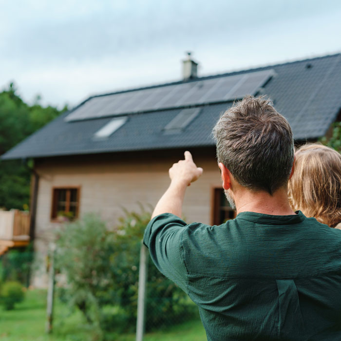 Rear view of dad holding daughter in arms and pointing to their house with solar panels